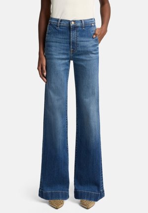 Personne portant un jean en denim bleu taille haute à jambes larges et des chaussures à bouts pointus cloutées, debout avec une main dans la poche.