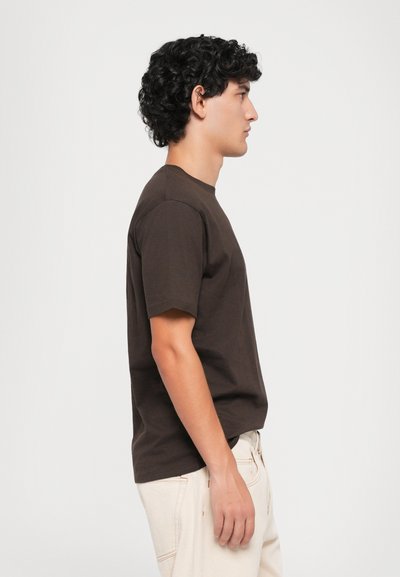 Jeune homme aux cheveux bouclés portant un t-shirt brun foncé et un pantalon beige clair, se tenant de côté devant un fond blanc uni.