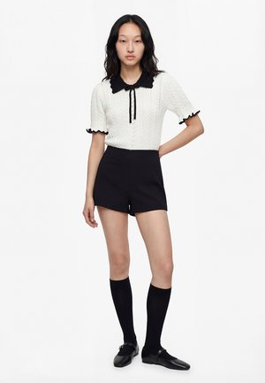 Junge Frau trägt ein weißes, kurzärmeliges Strickoberteil mit schwarzem Kragen, schwarze Shorts mit hoher Taille, kniehohe Socken und schwarze Ballerinas.