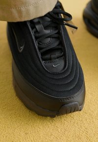 Sneaker noire Nike Air Max 97 avec tissu texturé, accents lisses, bout rond et branding subtil. La chaussure est partiellement couverte par une jambe de pantalon beige.