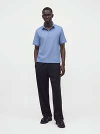 Homme debout portant un polo bleu clair à manches courtes, un pantalon noir et des chaussures noires sur un fond blanc uni.
