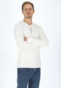 Vit långärmad henley-tröja med rundad hals och fyra bruna knappar. Ribbad textur, muddar med tumhål, ihop med blå jeans.