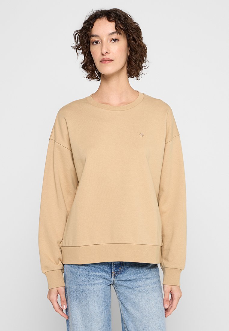 Sudadera de algodón beige con escote redondo, hombros caídos, puños de canalé y un lindo logo en el pecho, combinada con jeans de color azul claro.