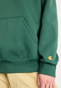 Vihreä collegepaita kengurutaskulla, resorilla varustetut hihansuut ja kultainen logokoriste oikeassa hihassa. Pehmeä pinta, rento malli.