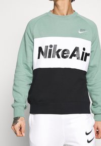 Grön, vit och svart sweatshirt med "Nike Air" i fet svart text. Tillverkad av mjukt tyg med ribbade ärmmuddar och rundad halsringning.