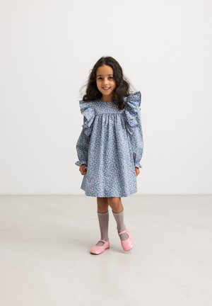 Jong meisje met lang krullend haar, een blauwe bloemenjurk, grijze kniekousen en roze schoenen, staand op een lichte vloer tegen een witte muur.