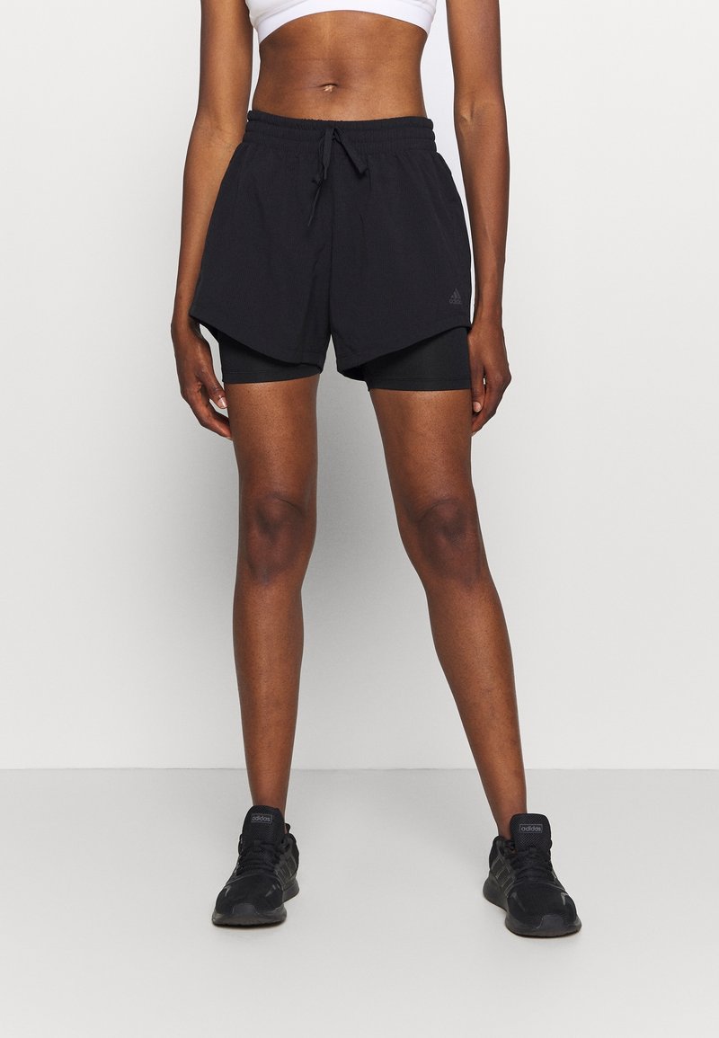 Zwarte sportshorts met een gevouwen ontwerp en een elastische tailleband, gecombineerd met zwarte compressieshorts en zwarte hardloopschoenen.