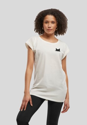 Femme aux cheveux bouclés portant un t-shirt blanc avec un petit motif de chat noir près de l'épaule gauche et des leggings noirs.