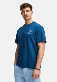 Camiseta de algodón azul con mangas cortas, escote redondeado y el texto "libera tu mente" bordado en azul claro en el lado izquierdo del pecho.
