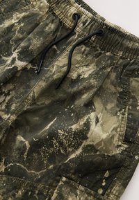 Green and beige camouflage shorts with an elastic waistband, drawstring closure, and pockets. Features a textured, splattered pattern.