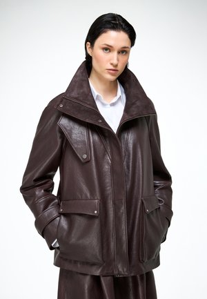 Vrouw met donker haar die een donkerbruin oversized leren jack draagt over een wit overhemd met kraag, staand met handen in de zakken.