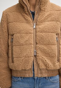 Beżowa polarowa kurtka z zamkiem z przodu, kieszeniami bocznymi, elastycznymi mankietami i prążkowanym dołem. Materiał ma miękką, teksturowaną powierzchnię.