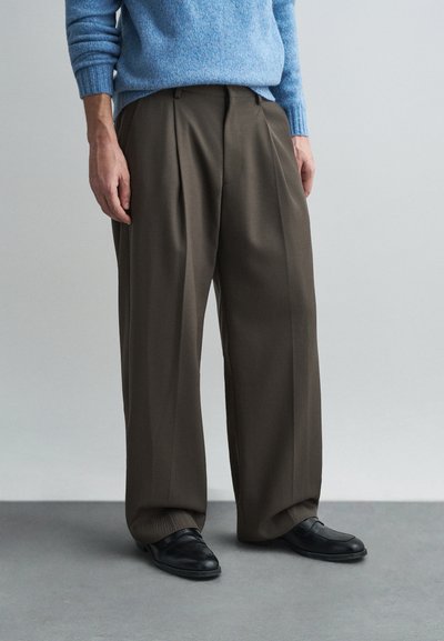 Pantalons plissés marron avec une coupe décontractée, en tissu lisse, associés à des chaussures noires à enfiler et un pull léger bleu.