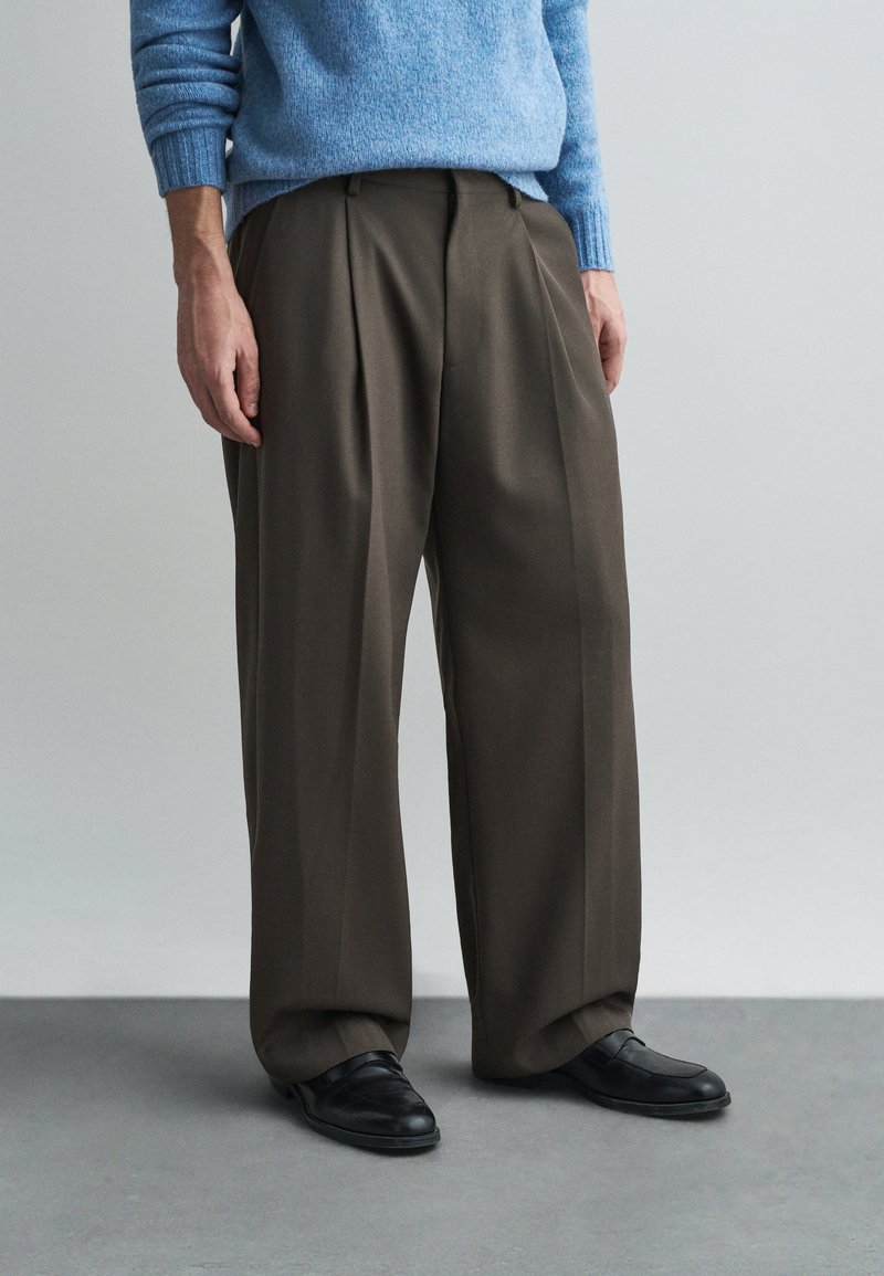 Barna, rakott szárú nadrág, laza fit, sima anyagból készült, fekete slip-on cipőkkel és világoskék pulóverrel párosítva.