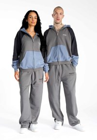Dvaja jednotlivci nosia zladené zip-up mikiny a tepláky v sivej a svetlomodrej farbe, ktoré majú vrecká, logo a nastaviteľné šnúrky.