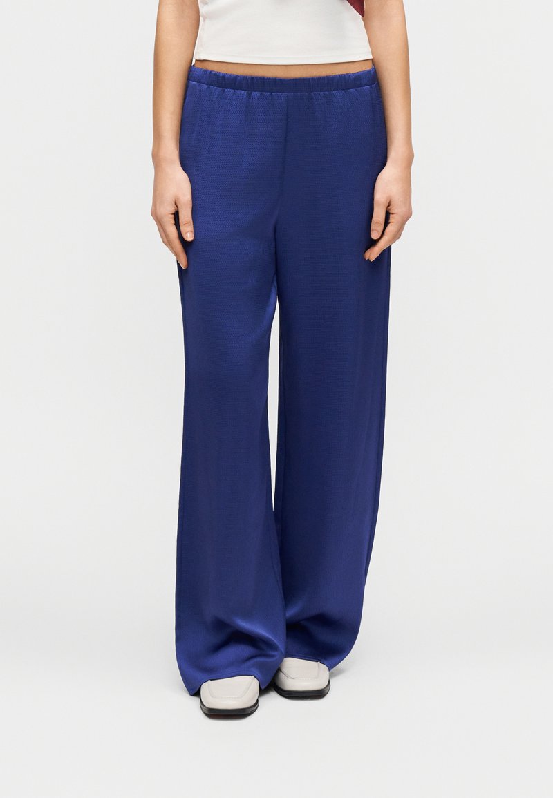 Φαρδιά, μπλε παντελόνια κατασκευασμένα από ένα λείο, ελαφρύ ύφασμα. Διαθέτουν ελαστική μέση και άνετη εφαρμογή. Διακοσμημένα με ελάχιστα ραφές.