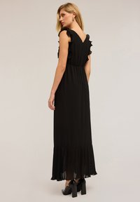 Langes schwarzes Kleid mit voluminösen Ärmeln, V-Ausschnitt am Rücken und plissiertem Saum. Kombiniert mit klobigen schwarzen Schuhen mit Absatz. Glatte Textur, elegante Silhouette.