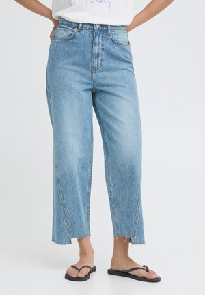 Žena v svetlomodrých širokých džínsových nohaviciach s oteplenými koncami a čiernych žabkách stojaca pred jednofarebným pozadím.