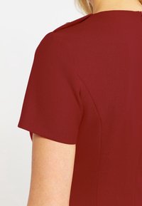 Abito bordeaux con maniche corte, tessuto texturizzato, silhouette aderente e chiusura con zip sul retro. Design elegante con sottili accenti sulle spalle.