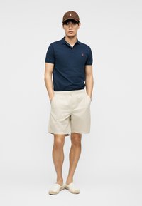 Mann trägt ein marineblaues Poloshirt, beige Shorts, braune Kappe und beige Slip-on-Schuhe, steht mit den Händen in den Taschen.