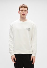 Κρεμ ζακέτα με στρογγυλή λαιμόκοψη και ribbed μανσέτες. Διαθέτει εκτύπωση μικρού μαύρου λογότυπου στο μπροστινό αριστερό μέρος. Συνδυάζεται με μαύρο παντελόνι.