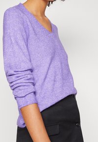 Pull lilas à col en V avec des manches retroussées, fabriqué dans un tissu doux et texturé. Associé à un bas noir ajusté pour un contraste.