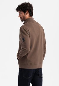 Bruine zip-up jas met een gestructureerd patroon op de mouwen, ribgebreide manchetten en een rechtopstaande kraag. Inclusief een logo op de mouw.