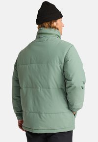 Giacca a vento verde chiaro con colletto alto, cuciture orizzontali e polsini elasticizzati, indossata con un cappello di maglia nero sulla schiena di una persona.