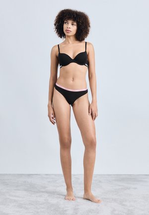 Sostén negro de tela suave y tirantes finos, combinado con bragas negras que tienen una cinturilla de marca rosa, modelo de pie sobre una superficie gris claro.