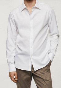 Chemise blanche à manches longues en tissu léger, dotée d'un col classique et d'une patte de boutonnage, associée à un pantalon marron.