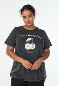 Frau mit lockigen Haaren, die ein dunkelgraues T-Shirt mit weißer Kirschen-Grafik und dem Schriftzug "The Cherry Edit Cherry vibes" trägt, die Hände locker verschränkt.