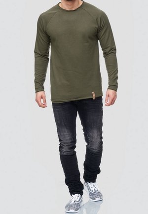 Homme portant une chemise à manches longues vert olive, un jean noir délavé et des baskets grises à motifs, debout devant un fond clair uni.
