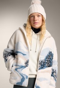 Femeie purtând o căciulă albă tricotată și o jachetă crem sherpa cu un model de peisaj montan albastru și negru, cu mâinile în buzunare.