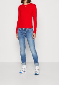 Maglietta rossa a maniche lunghe con scollatura rotonda, abbinata a jeans slim-fit blu chiaro, risvoltati alla caviglia. Sneakers bianche con dettagli blu.