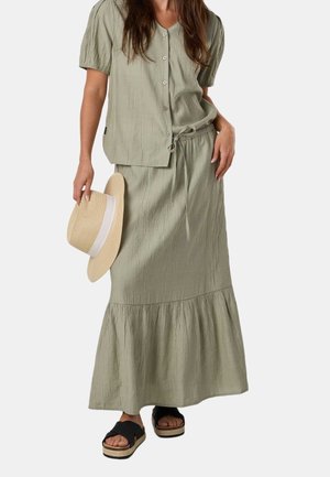 Frau trägt hellgrüne, geknöpfte Bluse und passenden langen Rock, hält beigefarbenen breitkrempigen Hut, trägt schwarze Plateausandalen.