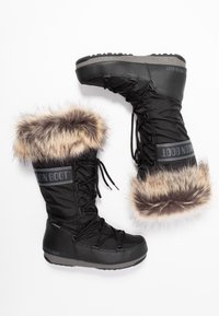 Botas de invierno negras con borde de piel de pelo, cordones en la parte delantera y suelas de goma gruesas, con la marca "Moon Boot" en el lateral y en la parte trasera.
