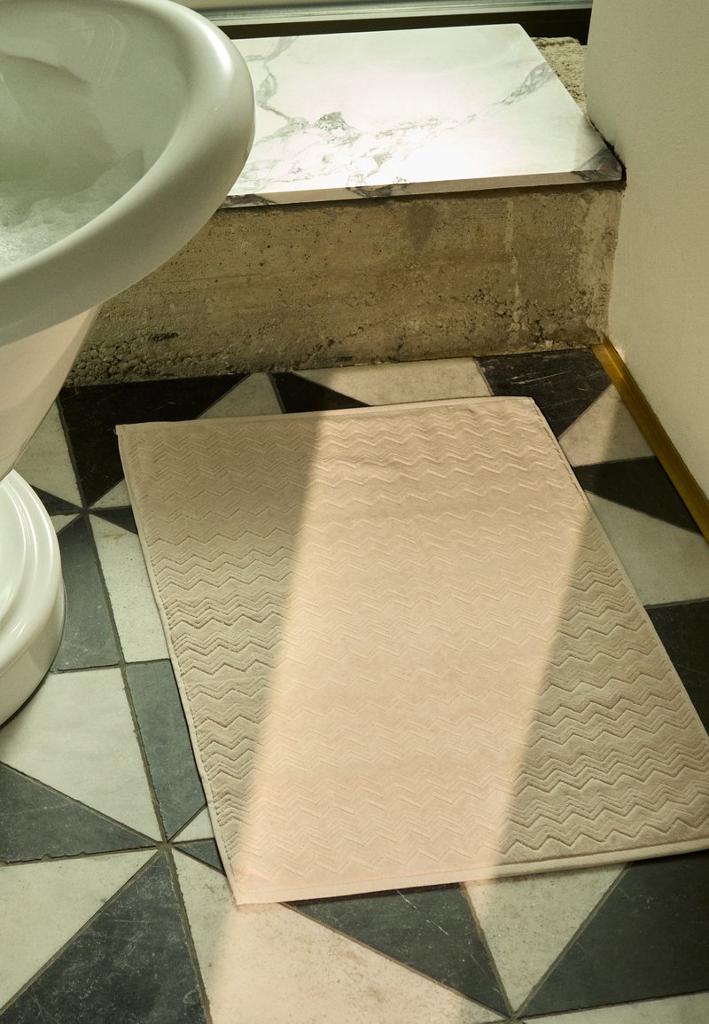 Tapete de banho em padrão zig-zag bege posicionado perto de uma pia pedestal branca, sobre um chão de azulejos preto e branco em padrão. A luz do sol forte projeta uma sombra.