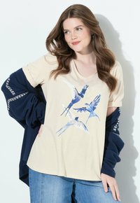 Mujer joven con cabello largo y castaño que lleva una camisa beige con estampado de pájaros azules, jeans azules y un cárdigan navy sobre los brazos, contra una pared blanca.