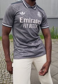 Mężczyzna w szarej koszulce piłkarskiej Real Madrid z logo "Emirates Fly Better", stojący na zewnątrz na kamiennej nawierzchni w pobliżu roślin i budynku.