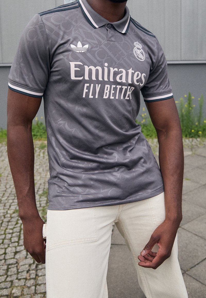 Mężczyzna w szarej koszulce piłkarskiej Real Madrid z logo "Emirates Fly Better", stojący na zewnątrz na kamiennej nawierzchni w pobliżu roślin i budynku.