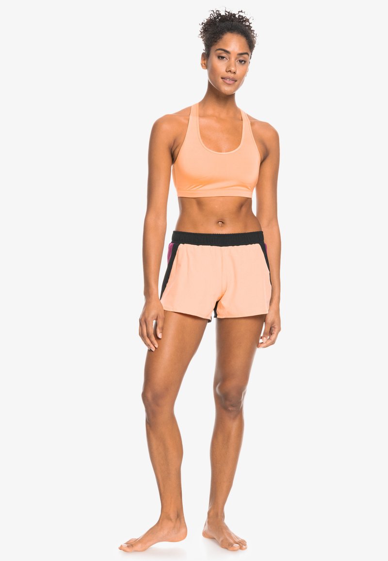 Sportbeha in licht perzik met een ronde halslijn, gecombineerd met perzik shorts met een zwarte elastische tailleband en zijaccenten.