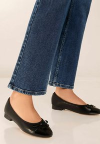 Schwarze flache Schuhe mit einer Schleifenapplikation, Lackzehen und mattem Obermaterial; kombiniert mit blauen Jeans aus Denim, die ein gerades Bein und einen umgeschlagenen Saum haben.