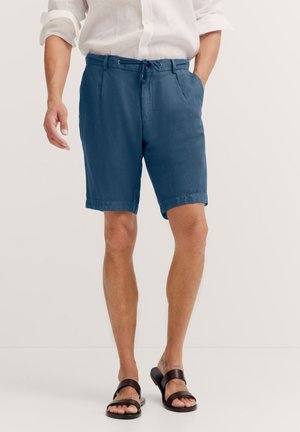 Herren blaue Leinenhosen mit elastischem Bund, seitlichen Taschen und einer lockeren Passform, kombiniert mit braunen Sandalen. Glatte Textur.