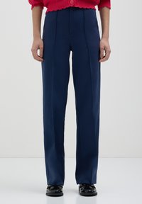 Personne portant un pantalon bleu marine à coupe droite, des mocassins noirs et un pull en laine rouge avec un gros bouton à la taille.