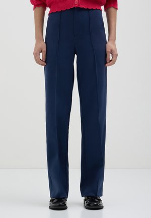 Personne portant un pantalon bleu marine à coupe droite, des mocassins noirs et un pull en laine rouge avec un gros bouton à la taille.