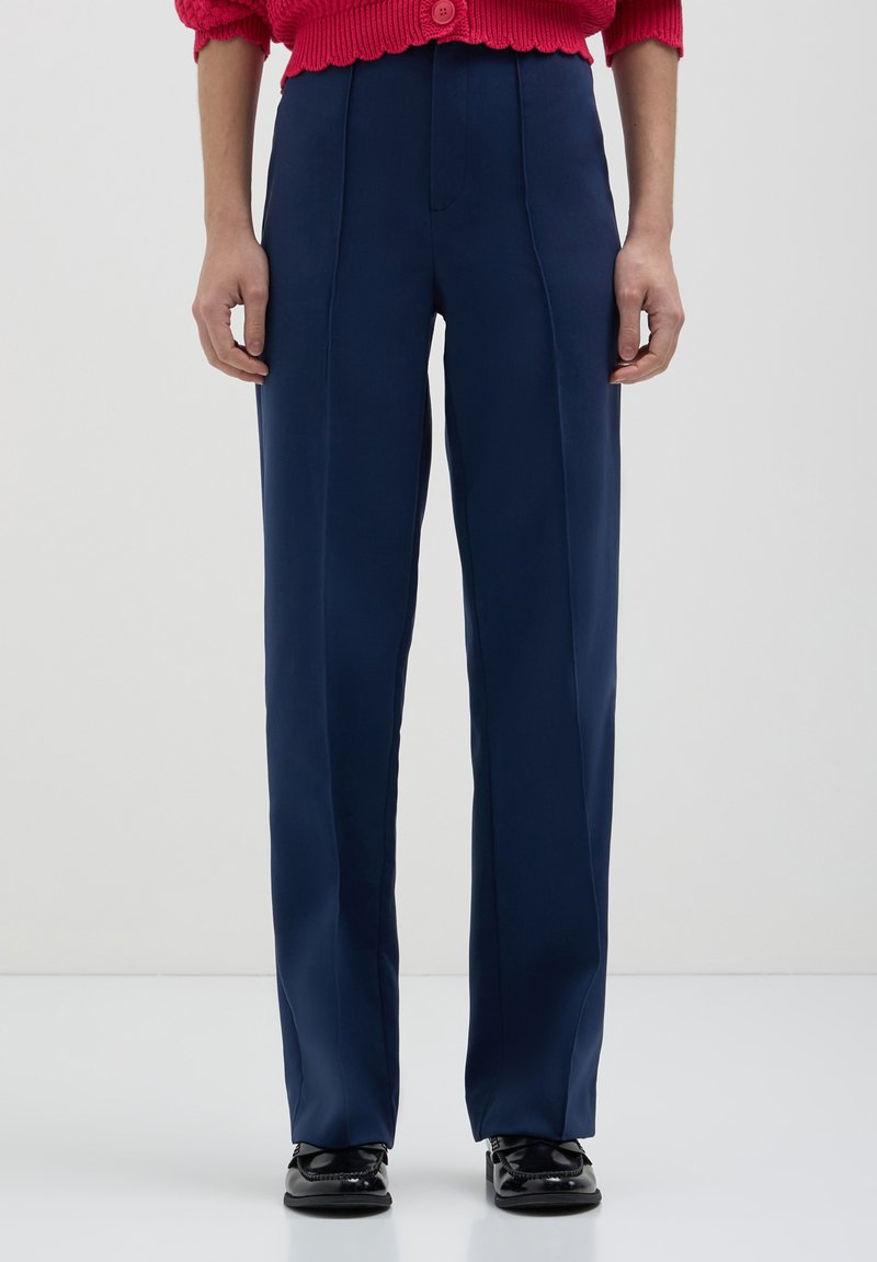 Personne portant un pantalon bleu marine à coupe droite, des mocassins noirs et un pull en laine rouge avec un gros bouton à la taille.