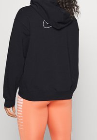 Felpa nera con cappuccio, dotata di tasca a marsupio frontale e polsini a costine, abbinata a leggings arancioni con strisce verticali bianche.
