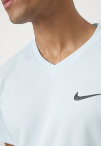 Hellblauses Sporthemd mit V-Ausschnitt, aus strukturiertem Stoff, mit einem schwarzen Nike-Logo auf der linken Seite.