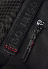 Black fabric backpack section featuring a textured mesh, embossed "HUGO" branding, and red stitching on the zipper pull cord.