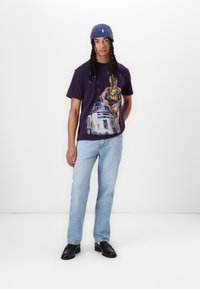 Lilla grafisk T-shirt med C-3PO og R2-D2, parret med lyseblå jeans og sorte loafers. Modellen bærer en blå ribbet hue.